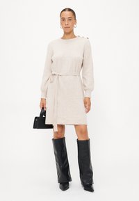 Beige gebreide jurk met een strikriem, lange pofmouwen en knoopaccenten op de schouders. Gepaarde met zwarte leren laarzen tot de knie.