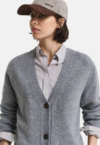 Grauer Wollcardigan mit V-Ausschnitt, ausgestattet mit einer Knopfleiste und gerippten Bündchen. Kombiniert mit einem gestreiften Hemd mit Knopfleiste und einer grauen Mütze.