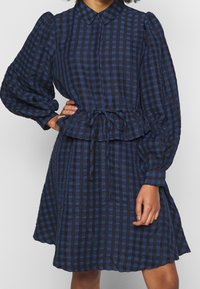 Sininen ja musta ruutuinen mekko, jossa on kaulus, puffatut hihat, vyötäröltä kiristetty muotoilu ja peplum-helmaversio. Pehmeä, teksturoitu kangas.