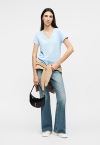 Γυναίκα που φοράει ανοιχτό μπλε T-shirt με V-λαιμόκοψη, μπεζ πουλόβερ δεμένο γύρω από τη μέση, τζιν με φαρδιά παντελόνια, λευκά αθλητικά παπούτσια, κρατώντας μαύρη τσάντα χειρός.