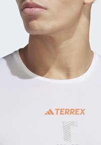 Primer plano del cuello y la parte inferior de la cara de una persona que lleva una camisa blanca con el logo en naranja "TERREX" y una letra "T" de rayas debajo.