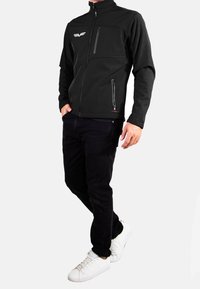 Veste softshell noire avec une fermeture éclair à l'avant, un col montant et deux poches latérales zippées. Présente un logo blanc sur la poitrine.