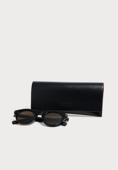 Lunettes de soleil ovales noires avec verres foncés placées devant un écrin rectangulaire en cuir noir Zegna sur fond blanc.