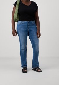 Svart kortärmad topp, blå raka jeans och svarta sandaler. Nyckeldetaljer inkluderar tygtextur och en grön vadderad väska.