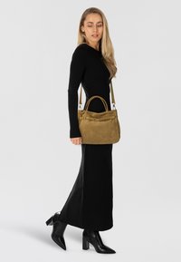 Borsa in pelle scamosciata verde oliva con doppio manico e tracolla, dotata di fibbia frontale e texture liscia, abbinata a un abito nero.