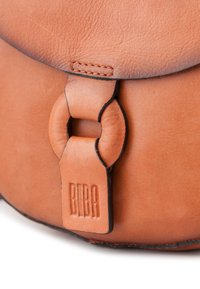 Bolso de cuero marrón claro con forma redondeada, que presenta una solapa con bordes negros y un cierre circular. "BIBA" grabado en la correa. Textura suave.