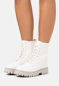 Valkoiset nauhakiinnitteiset nilkkasaappaat, joissa on teksturoitu nahkapinta, chunky harmaa ulkopohja ja pehmustettu kaulus.