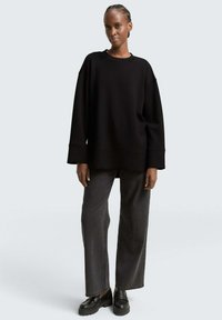 Vrouw in een losse zwarte trui, donkere wijde jeans en zwarte stevige loafers, staand tegen een effen lichtgrijze achtergrond.
