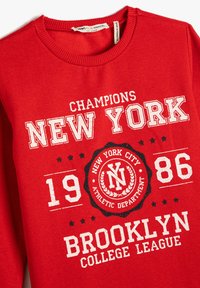 Czerwona bluza z białym napisem: "Mistrzowie Nowego Jorku 1986 Liga Brooklyn College" oraz okrągłym emblematem z napisem "Departament Sportowy Miasta Nowy Jork."