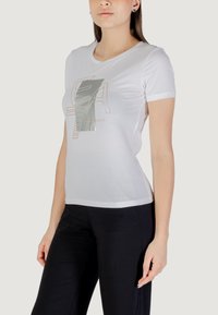 Camiseta blanca de manga corta con un diseño gráfico metálico en gris y beige, que presenta una forma ajustada y una textura suave de algodón.