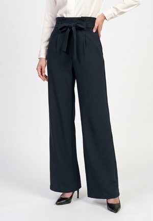 Femme portant un pantalon large taille haute bleu marine avec une ceinture nouée, associé à des talons noirs pointus et une blouse blanche.