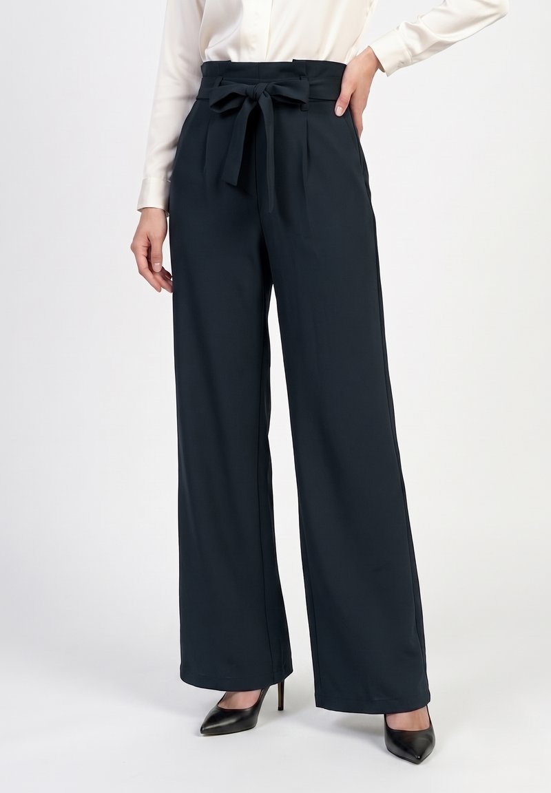 Femme portant un pantalon large taille haute bleu marine avec une ceinture nouée, associé à des talons noirs pointus et une blouse blanche.