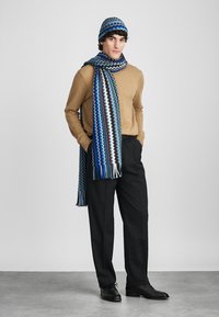 Άνδρας που φοράει μπλε καπέλο με σχέδιο πλεξούδας και μακριά ταιριαστή κασκόλ, καμήλικο πουλόβερ, μαύρο παντελόνι και μαύρα κοστούμια παπούτσια, στέκεται με τα χέρια στις τσέπες.