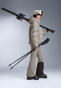 Kurtka s páperím s houndstooth vzorom, béžová a hnedá. Rozšírené nohavice s hnedým panelom, čierne lyže a palice. Biela lyžiarska prilba s clonou.