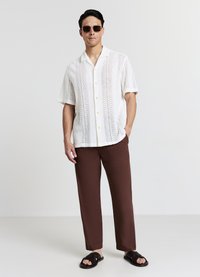 Weißes Kurzarmhemd mit Muster und Knöpfen, braune locker sitzende Hose und schwarze Sandalen, kombiniert mit Sonnenbrille.