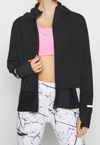 Donna che indossa una giacca nera con cappuccio sopra un reggiseno sportivo rosa e leggings bianchi con motivi a marmo nero, in piedi su uno sfondo semplice.