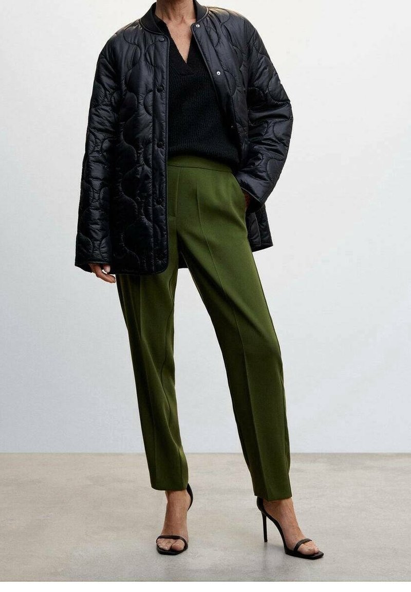 Personne portant une veste matelassée noire, un pull noir à col en V, un pantalon vert olive sur mesure et des sandales noires à talons, debout sur un sol gris.