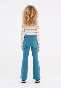 Pantaloni da sci blu con fondo svasato e tessuto flessibile, dotati di vita alta e cuciture a vista. Il modello indossa una maglietta a maniche lunghe con motivo.