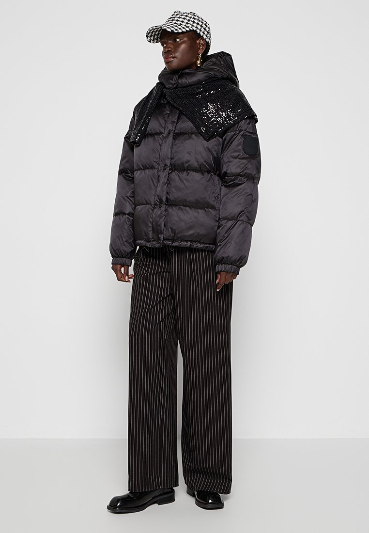 Schwarze Pufferjacke mit glänzender Oberfläche, oversized Schnitt, gerippte Bündchen, kombiniert mit schwarz-weißen gestreiften weiten Hosen und einer karierten Mütze.