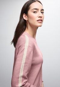Helles rosa Sweatshirt mit Rundhalsausschnitt und cremefarbenen Streifen an den Ärmeln. Die Textur wirkt weich und gestrickt und zeigt eine figurbetonte Silhouette.