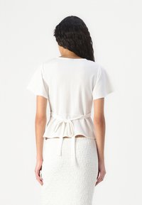 Hvit peplum-top med kortermede og bakbånddetalj. Lett stoff, glatt tekstur, med rund hals og utvidet søm.