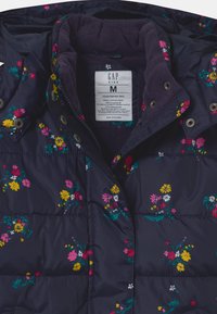 Giacca a piumino blu navy con motivo floreale in rosa, giallo e bianco. Presenta un colletto foderato in pile, chiusure a scatto e tessuto resistente all'acqua.