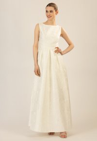 Une longue robe de mariée sans manches en crème avec une texture florale, une jupe plissée et un décolleté droit. Des sandales fines à talons argentés.