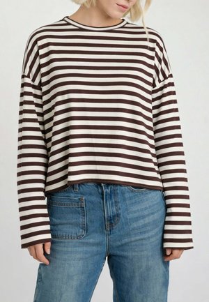 Persona che indossa una maglietta a maniche lunghe con strisce orizzontali marroni e bianche, infilata leggermente in un paio di jeans a vita alta, sbiaditi e di colore blu.