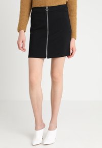 Černá mini sukně se zapínáním na zip vpředu a stříbrnými kovovými doplňky, vyrobená z přiléhavé, hladké látky. Nošena s bílými botami s otevřenou špičkou.