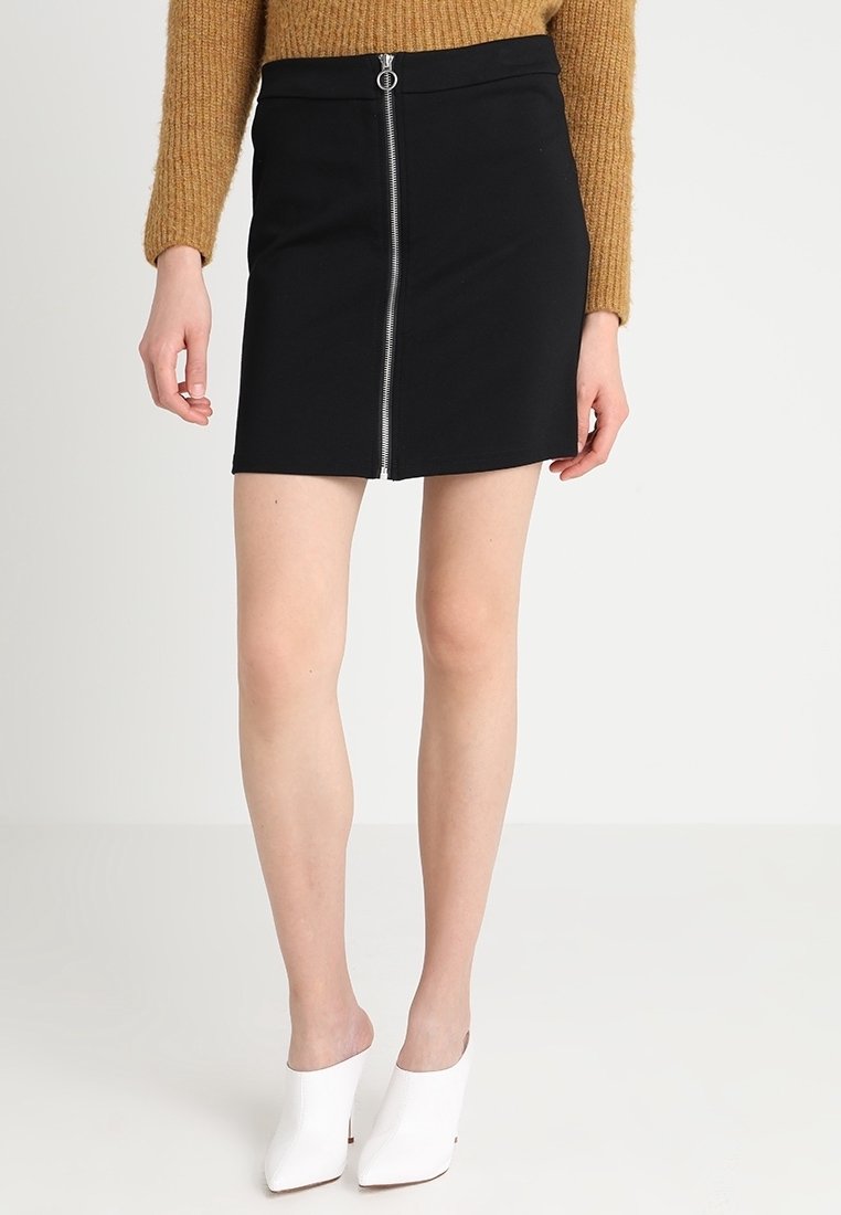 Černá mini sukně se zapínáním na zip vpředu a stříbrnými kovovými doplňky, vyrobená z přiléhavé, hladké látky. Nošena s bílými botami s otevřenou špičkou.