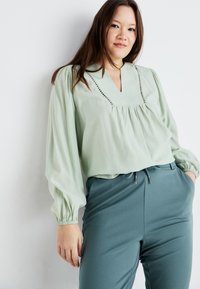 Světlezelená blůza s výstřihem do V, sklady na přední straně a dekoračním prošíváním. V kombinaci s tyrkysovými slim-fit kalhotami se šňůrkou.