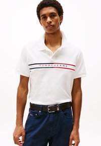 Camisa polo blanca con cuello, con franjas horizontales rojas y azul marino. En el frente se muestra el logo "TOMMY JEANS." Lleva un cinturón negro.