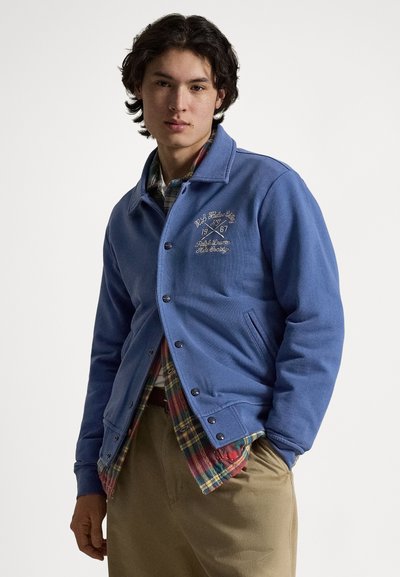 Plava dukserica s gumbima, izvezeni logotip, bočni džepovi i mekani materijal, slojevito nošena preko karirane košulje.