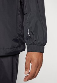Black waterproof jacket with elastic cuffs, textured fabric, and "DRYVENT" logo on sleeve. Hand resting on midsection, showing a close-up view.