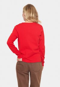 Rode lange mouwen top met een eenvoudige ronde halslijn. De stof is glad en nauwsluitend, gecombineerd met lichtbruine high-waisted broek.