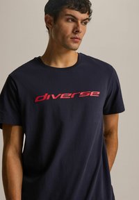 Granatowa koszulka bawełniana z wyraźnym różowym napisem "diverse" z przodu, z okrągłym dekoltem i krótkimi rękawami.