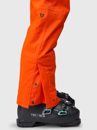 Lyse orange snepants med tryknapper over sorte skistøvler med spænder, vist fra siden på en grå baggrund.