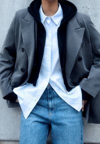 Blazer gris croisé porté sur un sweat à capuche noir et une chemise blanche à rayures, associé à un jean bleu clair. Tissus texturés et look superposé.