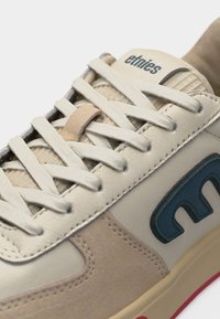 Κοντινό πλάνο σε παπούτσι τύπου sneaker σε μπεζ και κρεμ αποχρώσεις με λευκά κορδόνια, μπλε λογότυπο "etnies" στο πλάι και επένδυση στη γλώσσα με τη λέξη "etnies" σε σκούρα γράμματα.