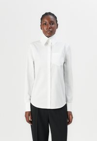 Weißes Hemd mit spitzem Kragen, langen Ärmeln und Brusttasche; glatte Textur und abgerundeter Saum; kombiniert mit schwarzen Hosen.