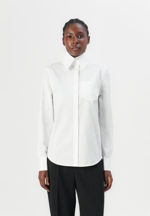 White button-up shirt with a pointed collar, long sleeves, and a chest pocket; smooth texture and rounded hem; paired with black trousers.