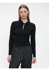 Schwarzes Langarmoberteil mit Knotenkollardetail und Schlüssellochöffnung; kombiniert mit grauen Nadelstreifenhose, die eine glatte Textur zeigt.