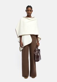 Biały poncho z brązowymi wykończeniami, zestawione z brązowymi spodniami o szerokich nogawkach oraz brązową torbą z srebrnymi akcentami. Gładki materiał, minimalistyczny design.