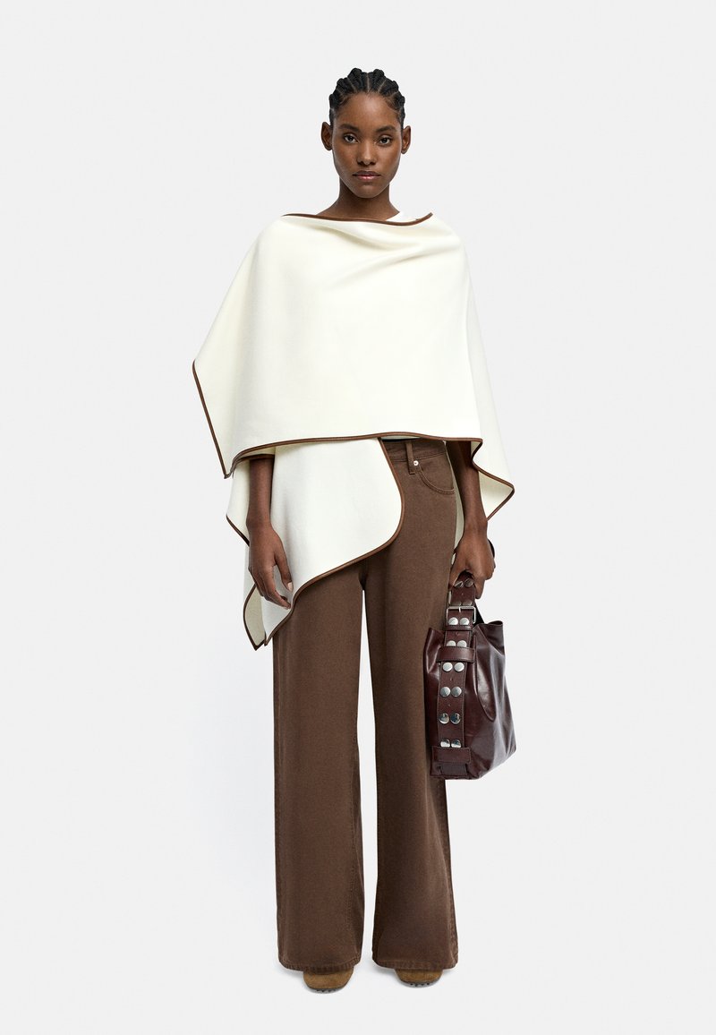 Biały poncho z brązowymi wykończeniami, zestawione z brązowymi spodniami o szerokich nogawkach oraz brązową torbą z srebrnymi akcentami. Gładki materiał, minimalistyczny design.