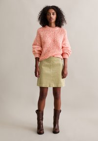 Gebreide oranje trui met een ronde hals en ballonmouwen, gecombineerd met een lichtgroene rok. Model draagt bruine leren laarzen.