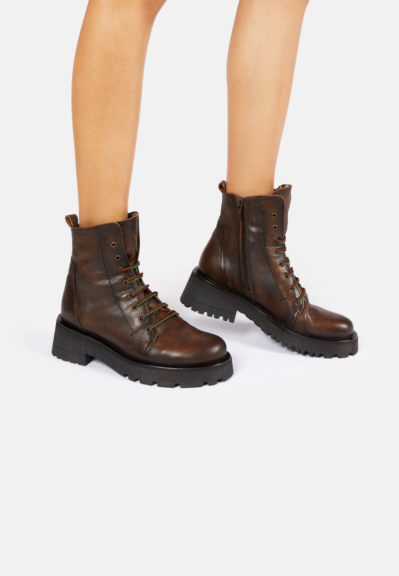 Botas de cuero marrón hasta el tobillo con puntera redonda, cierre de cordones en la parte frontal, detalles verdes, cremallera lateral y suela gruesa negra con dibujo.