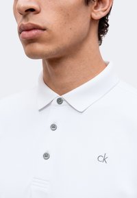 Gros plan sur une personne portant un polo blanc à trois boutons et un petit logo "ck" sur la poitrine.