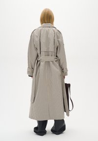 Trench coat a motivo check beige e marrone, con cintura in vita, maniche ampie e uno spacco sul retro. Abbinato a pantaloni neri e una borsa marrone.