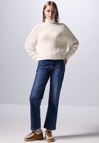 Cremefarbener, gerippter Strickrollkragenpullover mit breiten Bündchen, kombiniert mit blauen, geraden Jeans und braunen Schuhen mit Fellakzenten.