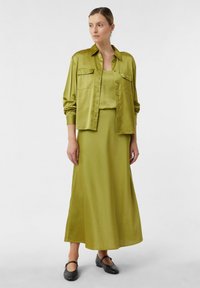 Vrouw in een glanzend olijfgroen satijnen shirt, bijpassend tanktop, lange rok en zwarte Mary Jane-schoenen, staand tegen een effen witte achtergrond.